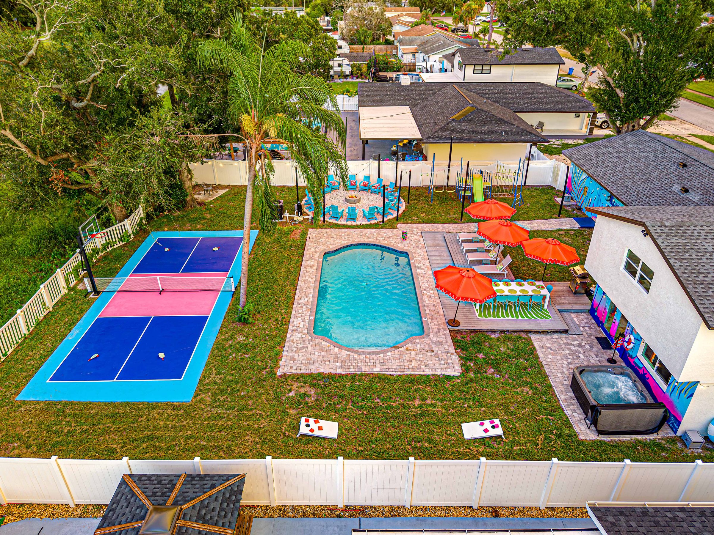 The whole backyard from above: private pickleball and basketball court, pool deck with umbrellas, hot tub, play set and fire pit